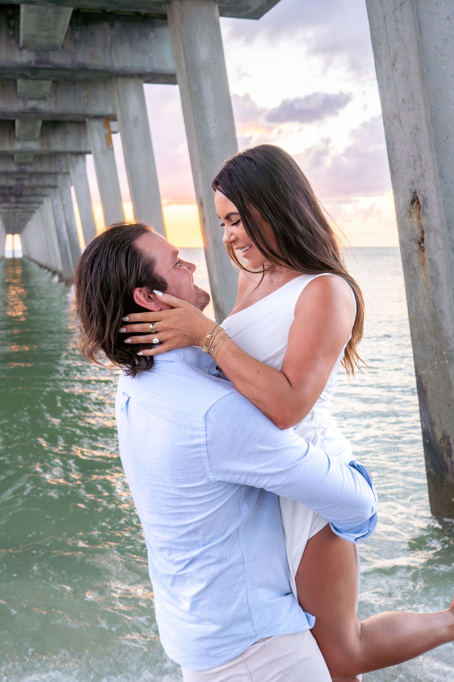 Romantic couple gazing at each other lovingly under a beach pier at colorful sunset.