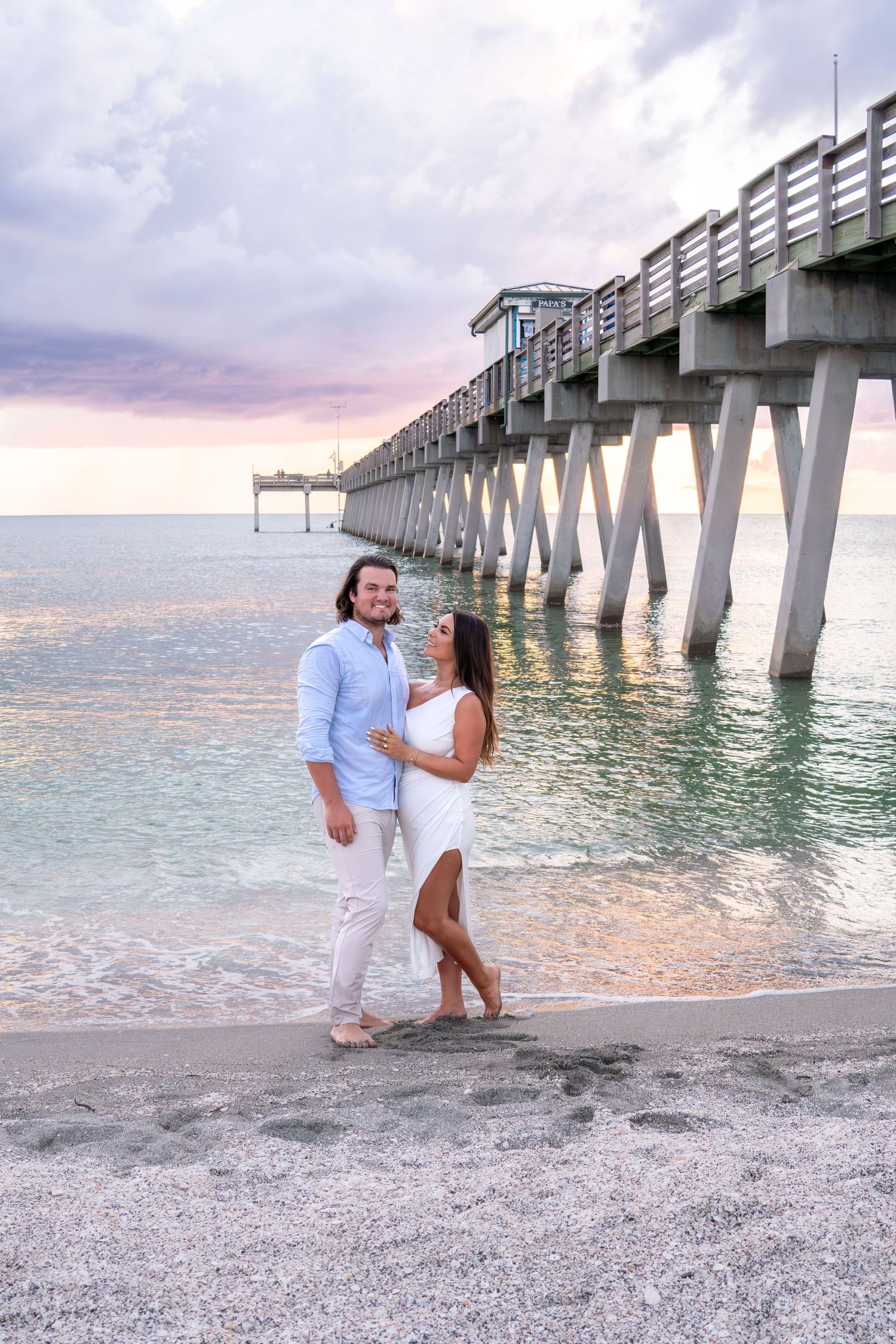 Happy engaged couple standing together on sandy beach beside a wooden pier at pastel sunset.
