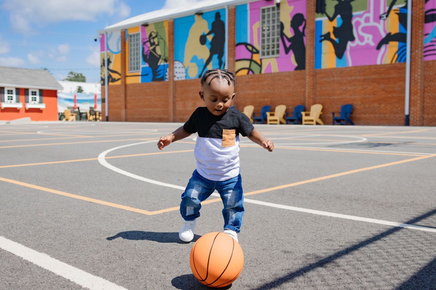 A young child plays with a basketball on an outdoor court with colorful murals in the background.