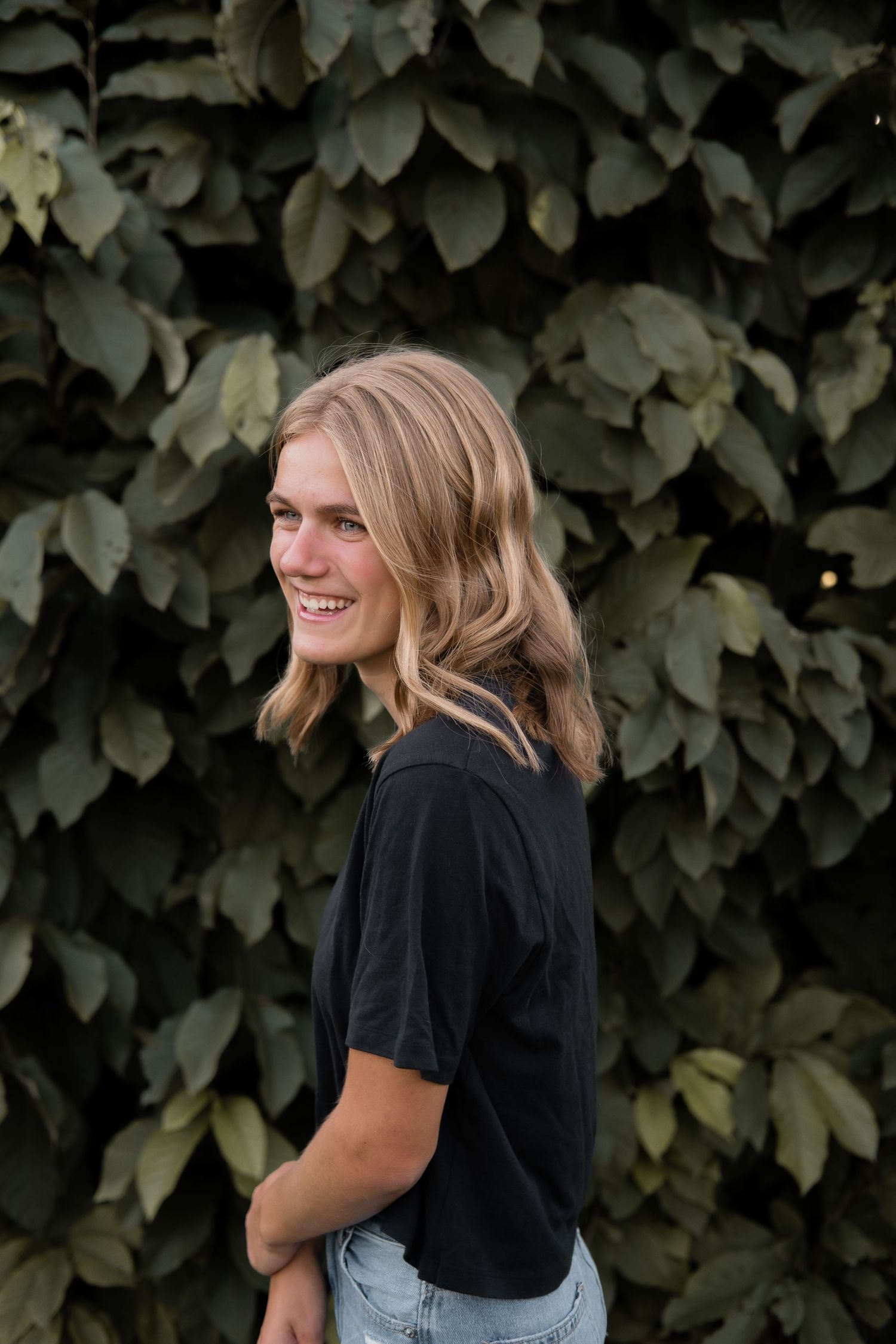 Someone in a black t-shirt poses casually against a backdrop of lush green leaves.