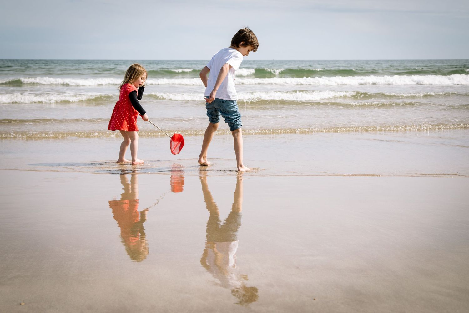 Two figures playing at the water's edge on a beach with their reflections in the wet sand.