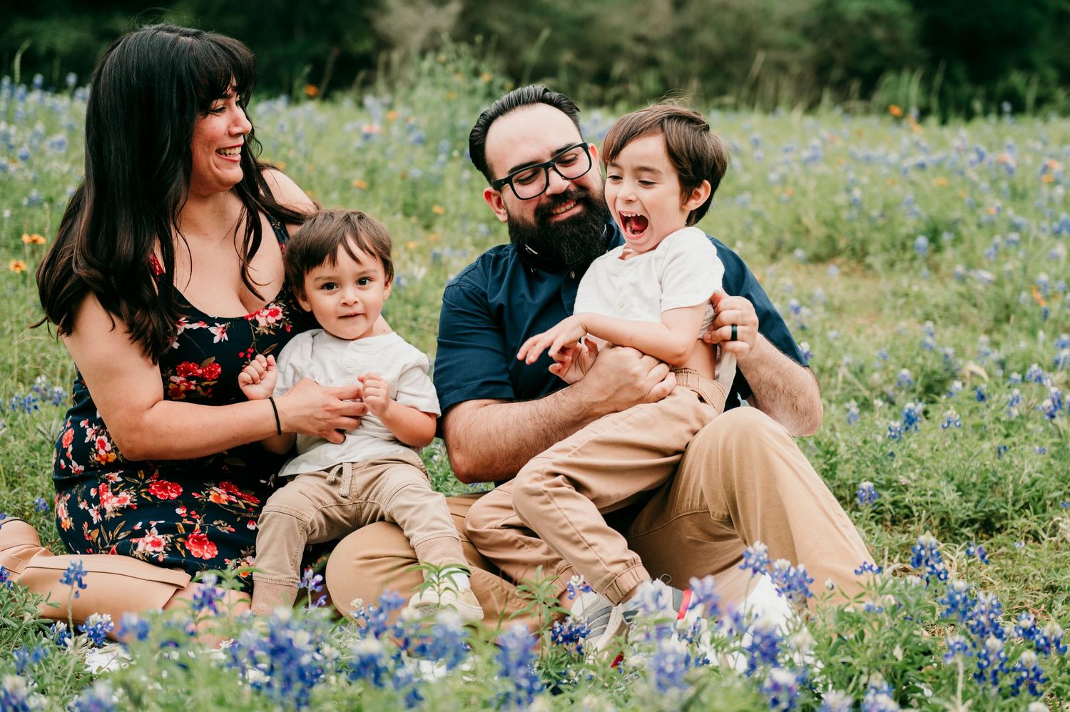 A mother, father, and two boys sitting in a field of bluebonnets laughing and playing together