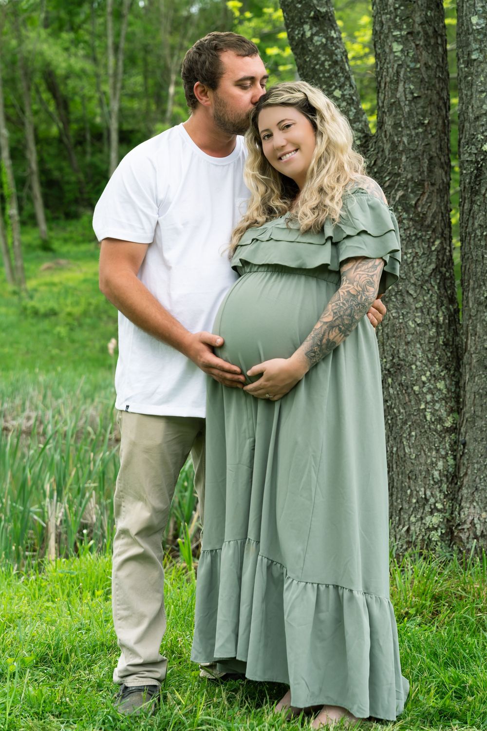 A couple embraces outdoors in a forest setting, wearing casual attire with a sage green maxi dress against tree trunks.