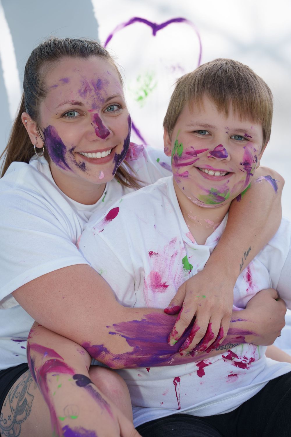 A smiling woman and boy covered in colorful purple and pink paint on their faces and white shirts, posing together.
