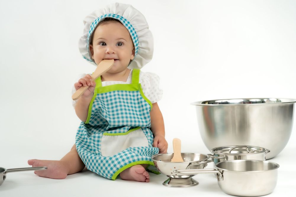 Tiny chef wearing blue gingham and white hat sits near stainless steel mixing bowls.