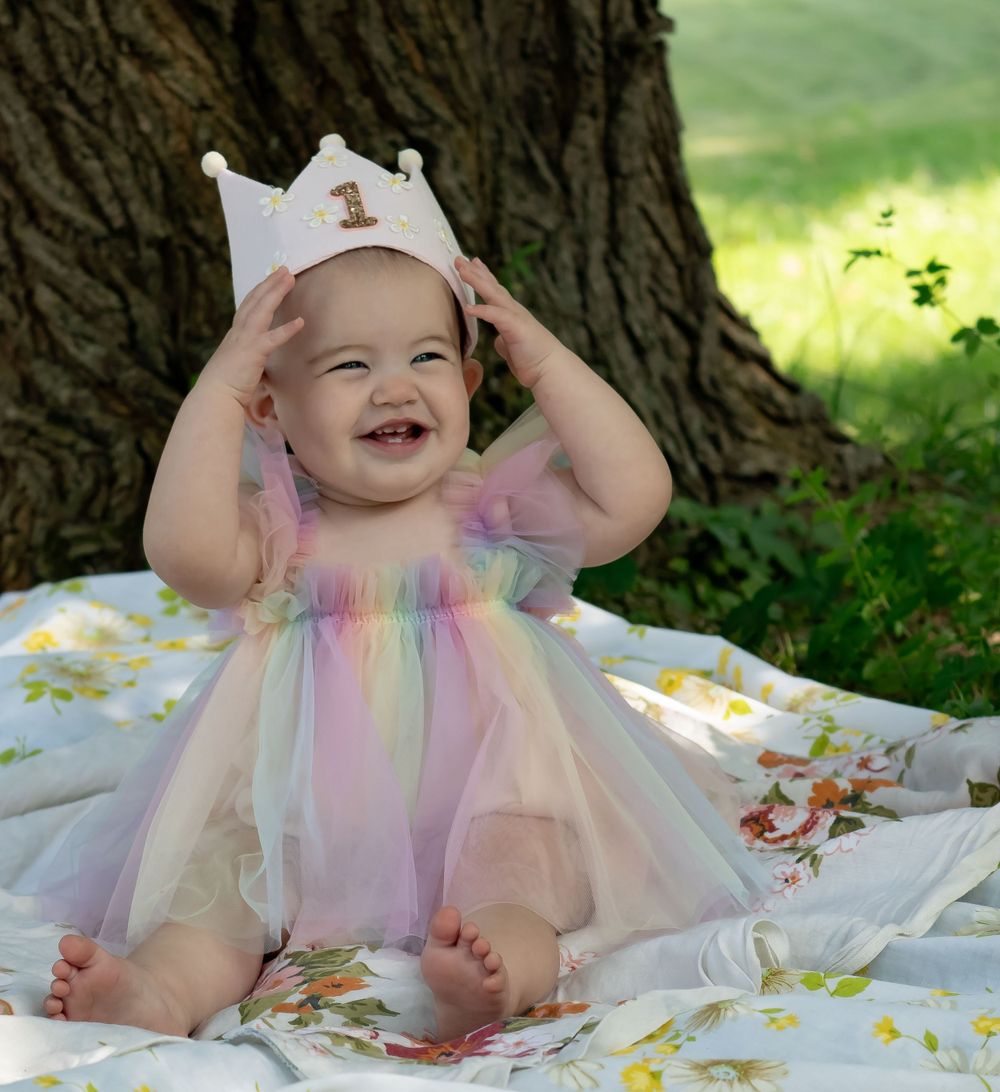A festive birthday setup featuring a pastel rainbow tulle dress and gold crown on a floral blanket.