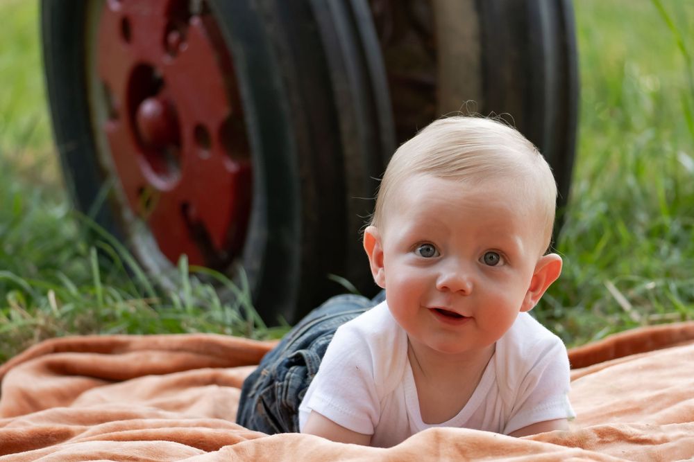 Baby smiling while lying on orange blanket with vintage tractor in background.