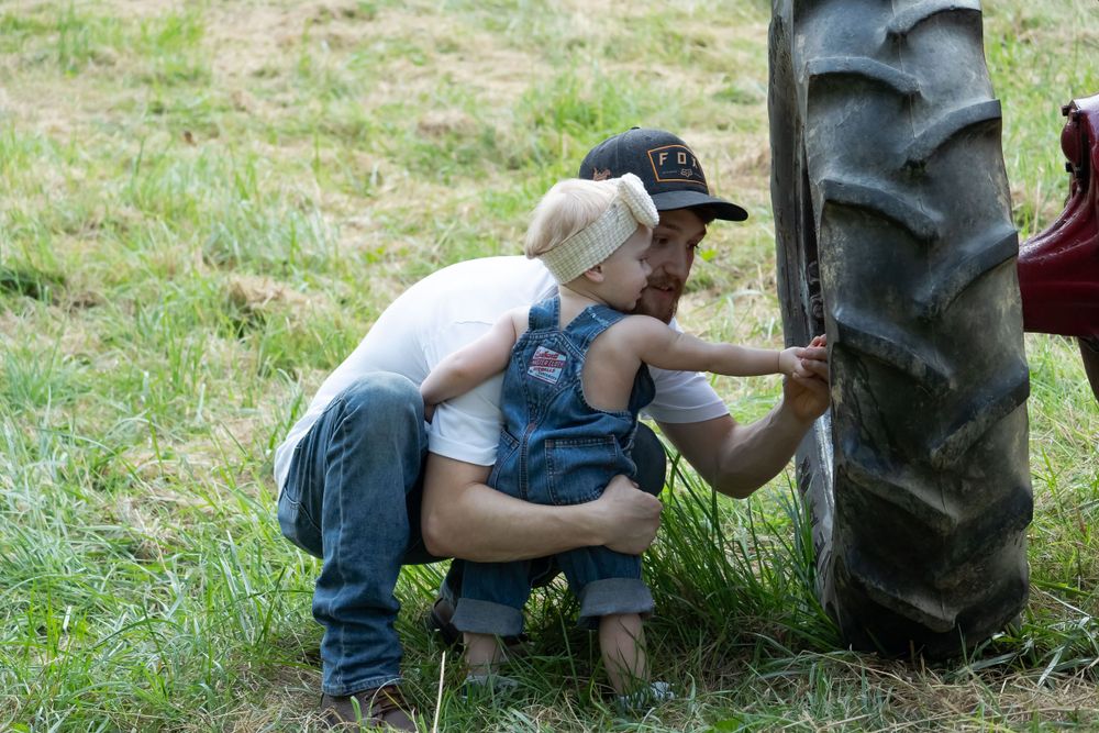 Small child in overalls crawls near large tractor tires in grassy field.