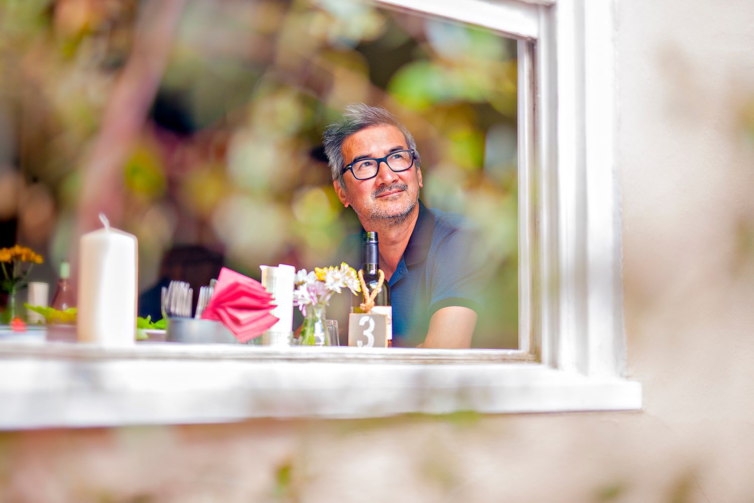 A candid view through a window of someone dining at a table with flowers and glasses.