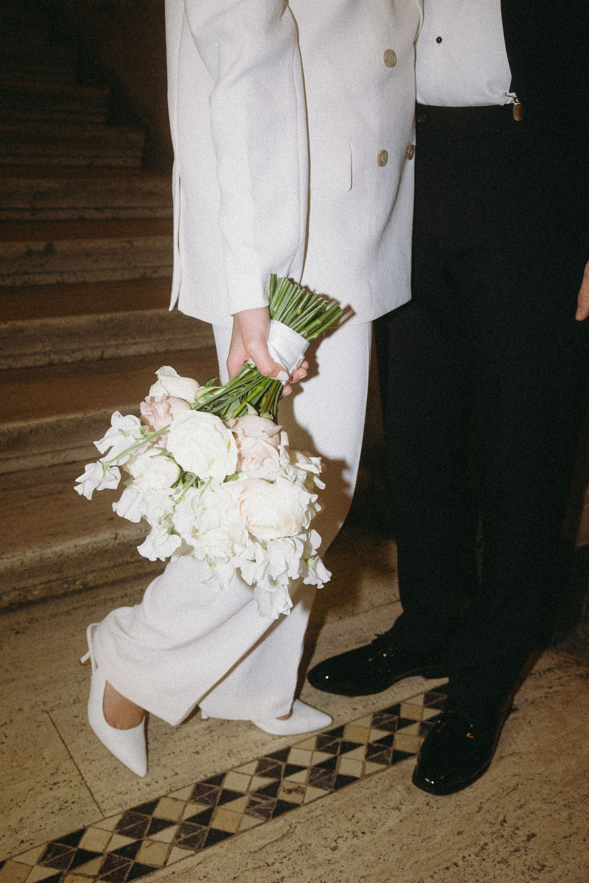 Bride in white blazer holding white peony bouquet standing beside groom on steps.