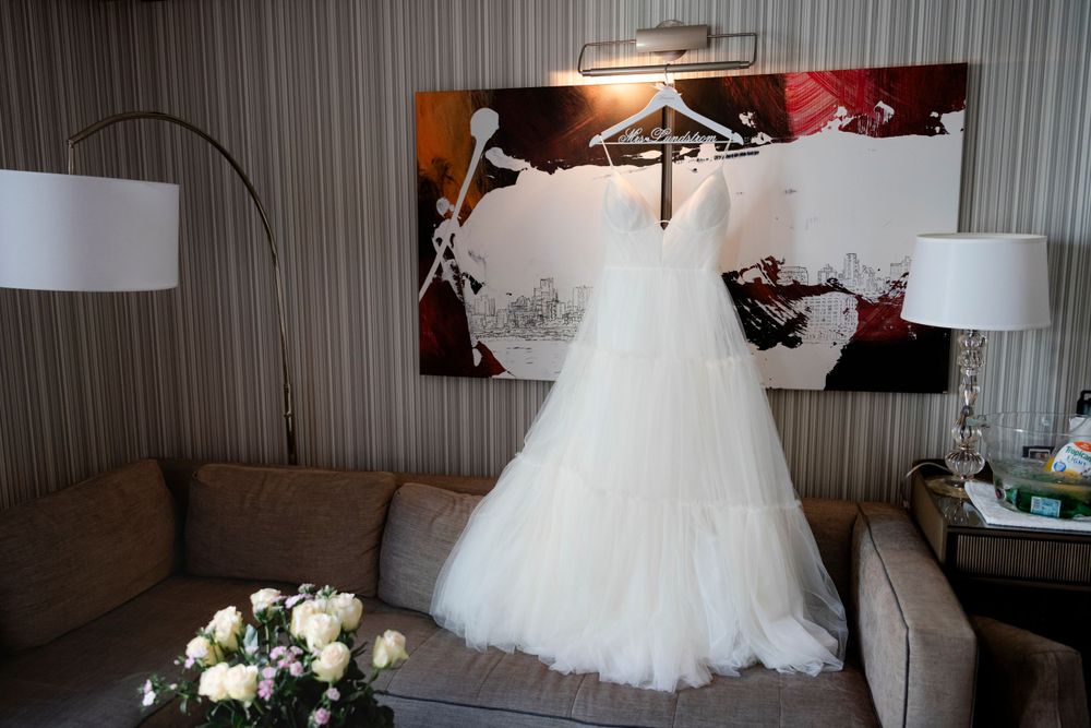 A white tulle wedding dress hangs on display above a gray couch with floral bouquet and table lamps in a hotel room.