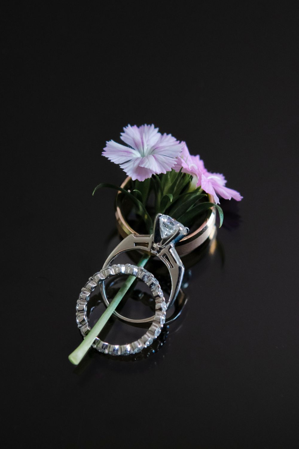 Diamond engagement ring and wedding band with pink dianthus flowers on a dark background.