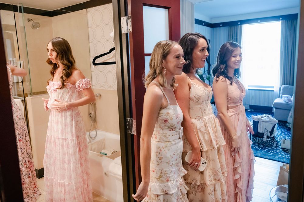 Multiple attendants in pink and white floral dresses stand together in a hotel room before an event.