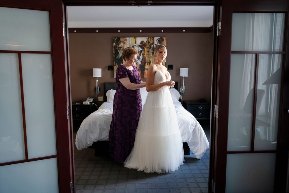 Two figures in elegant gowns prepare in a hotel room with a white bed visible in the background.