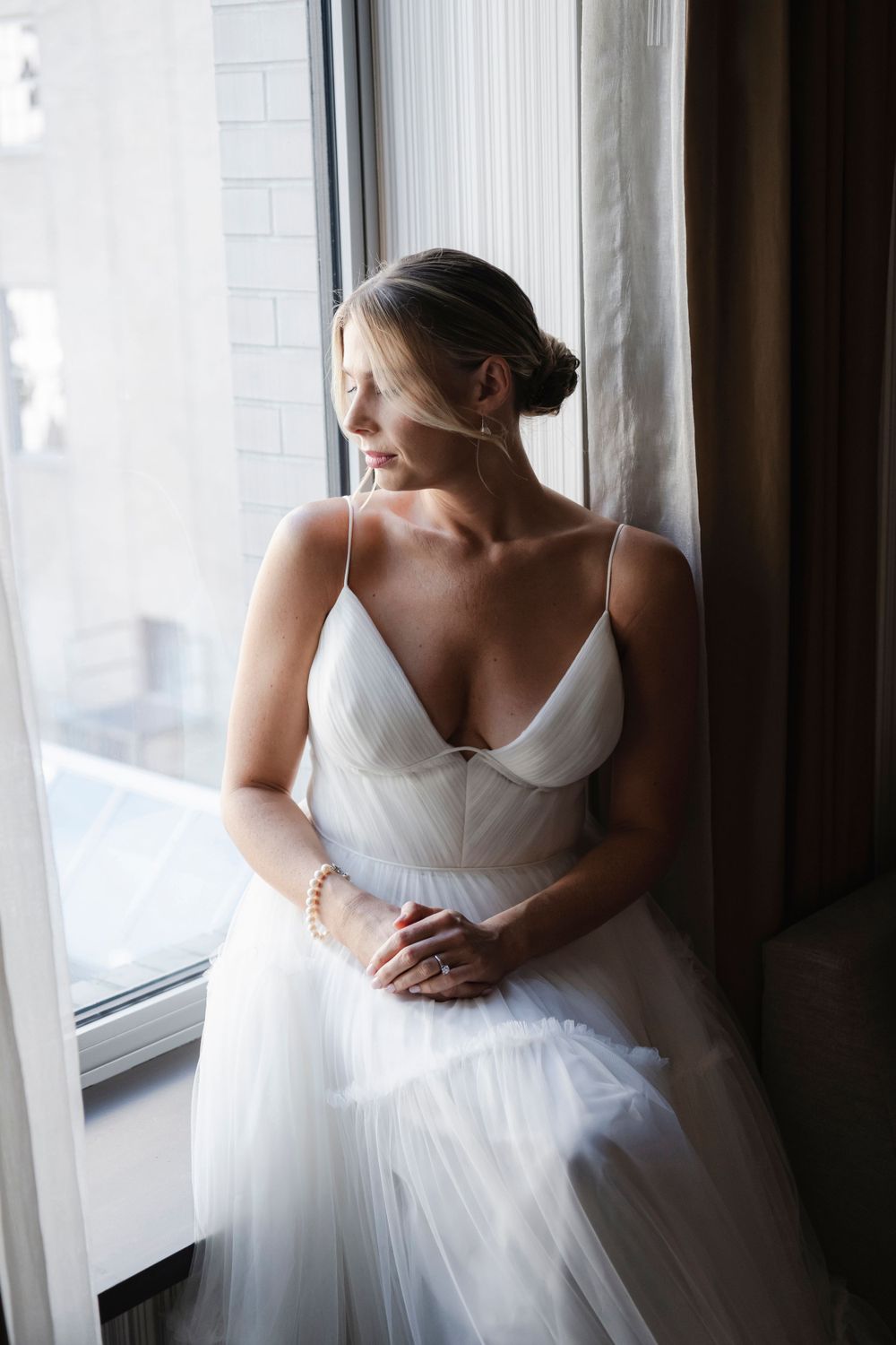 A person in an elegant white wedding gown with spaghetti straps gazes pensively through a window draped with sheer curtains.