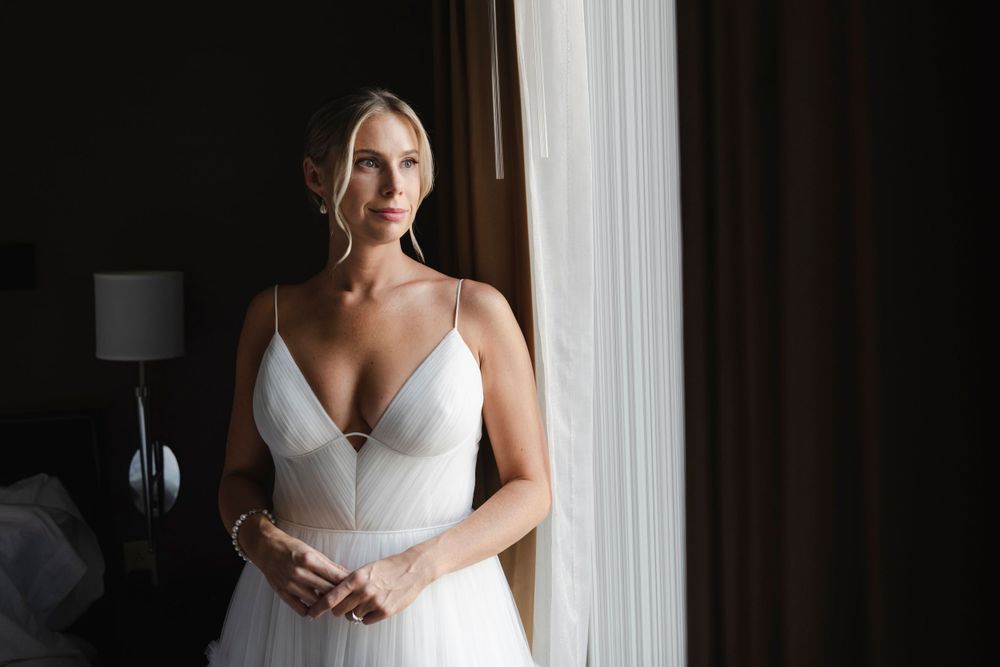 A bride in a sleeveless white gown stands elegantly by a window with natural light streaming in.