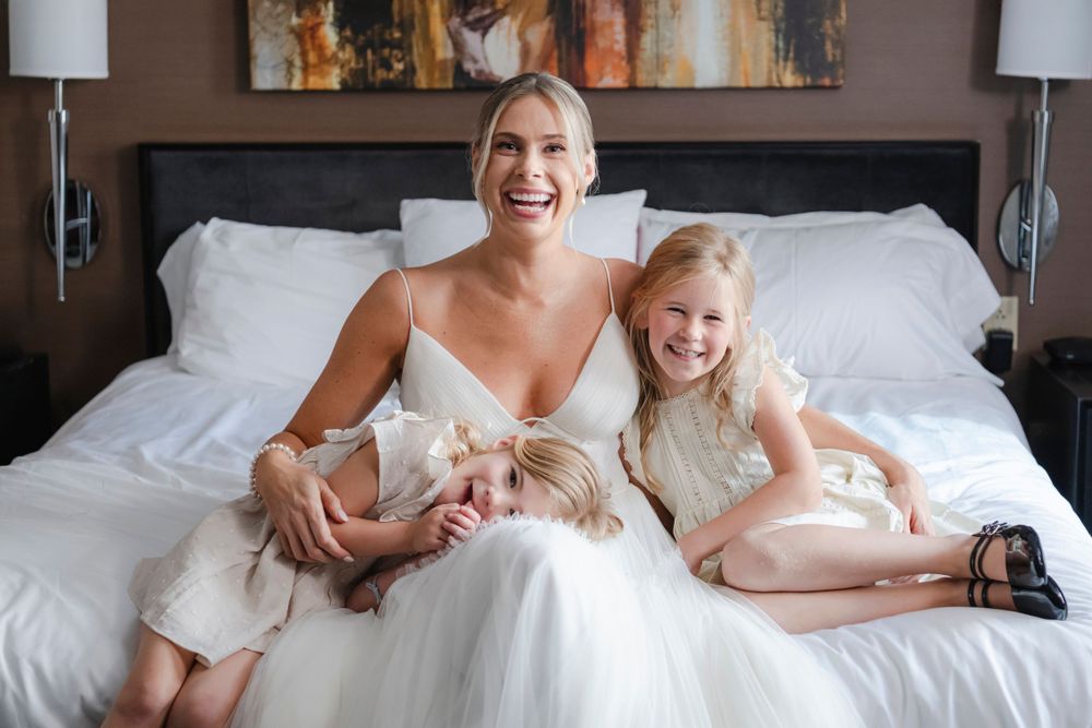 A bride and two flower girls in white outfits sit together on a bed with white linens, sharing joyful laughter and smiles.
