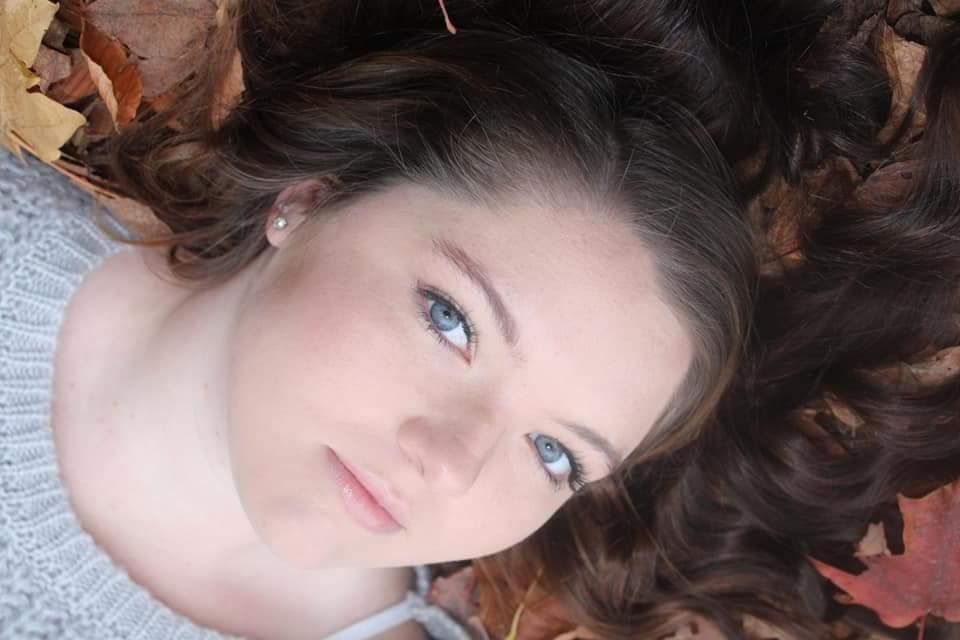 Young woman with blue eyes and curly brown hair lying among colorful autumn leaves outdoors.
