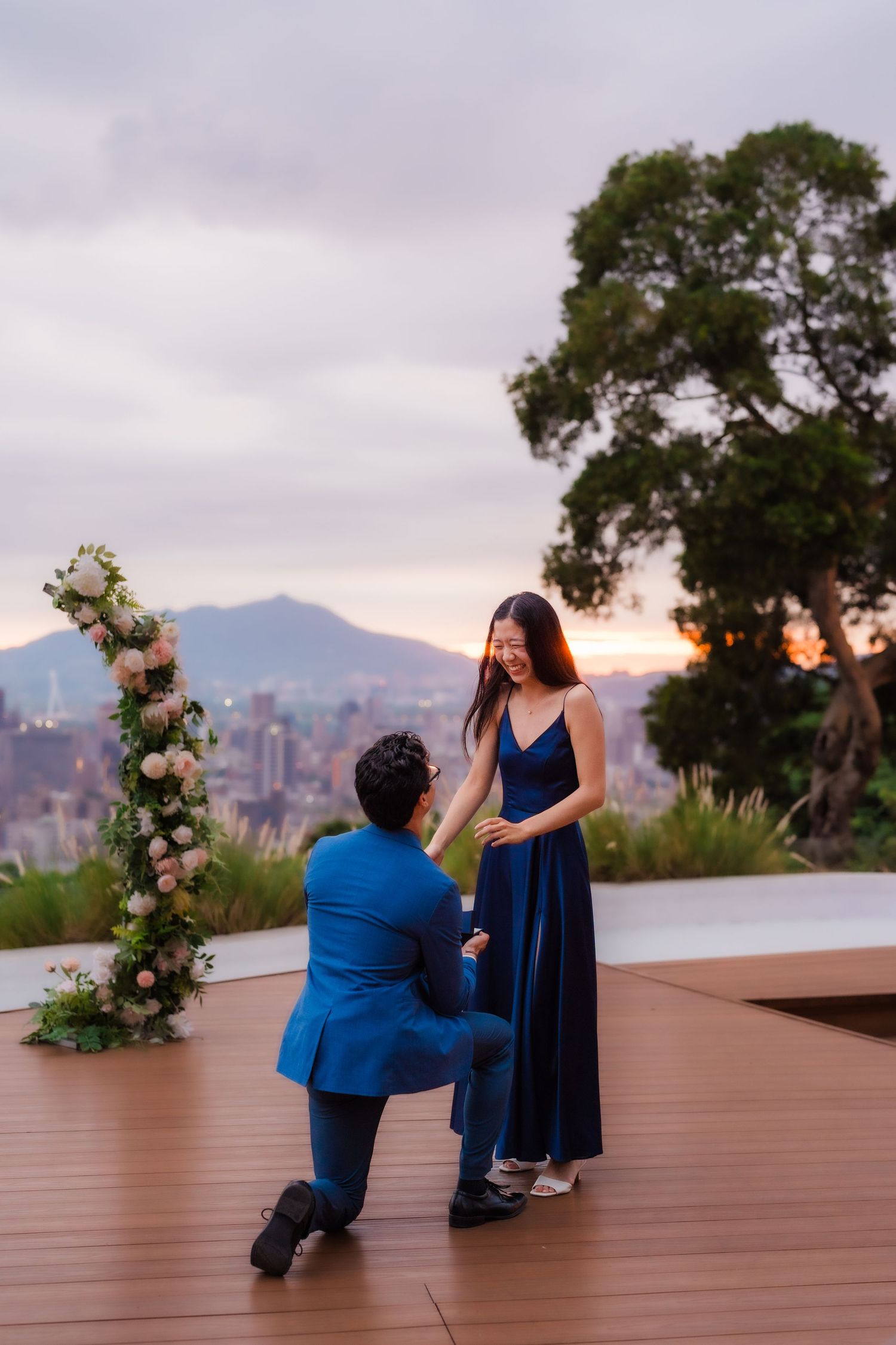 Marriage proposal at sunset with floral arch and city skyline backdrop.