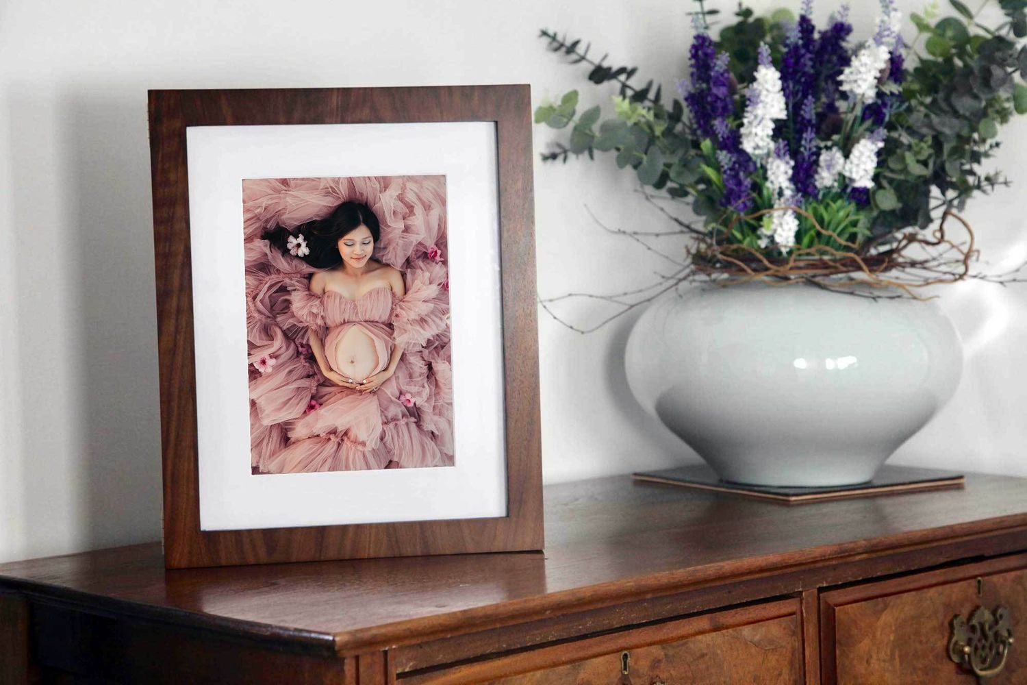 Framed maternity photo displayed with purple flowers in white vase on wooden furniture.