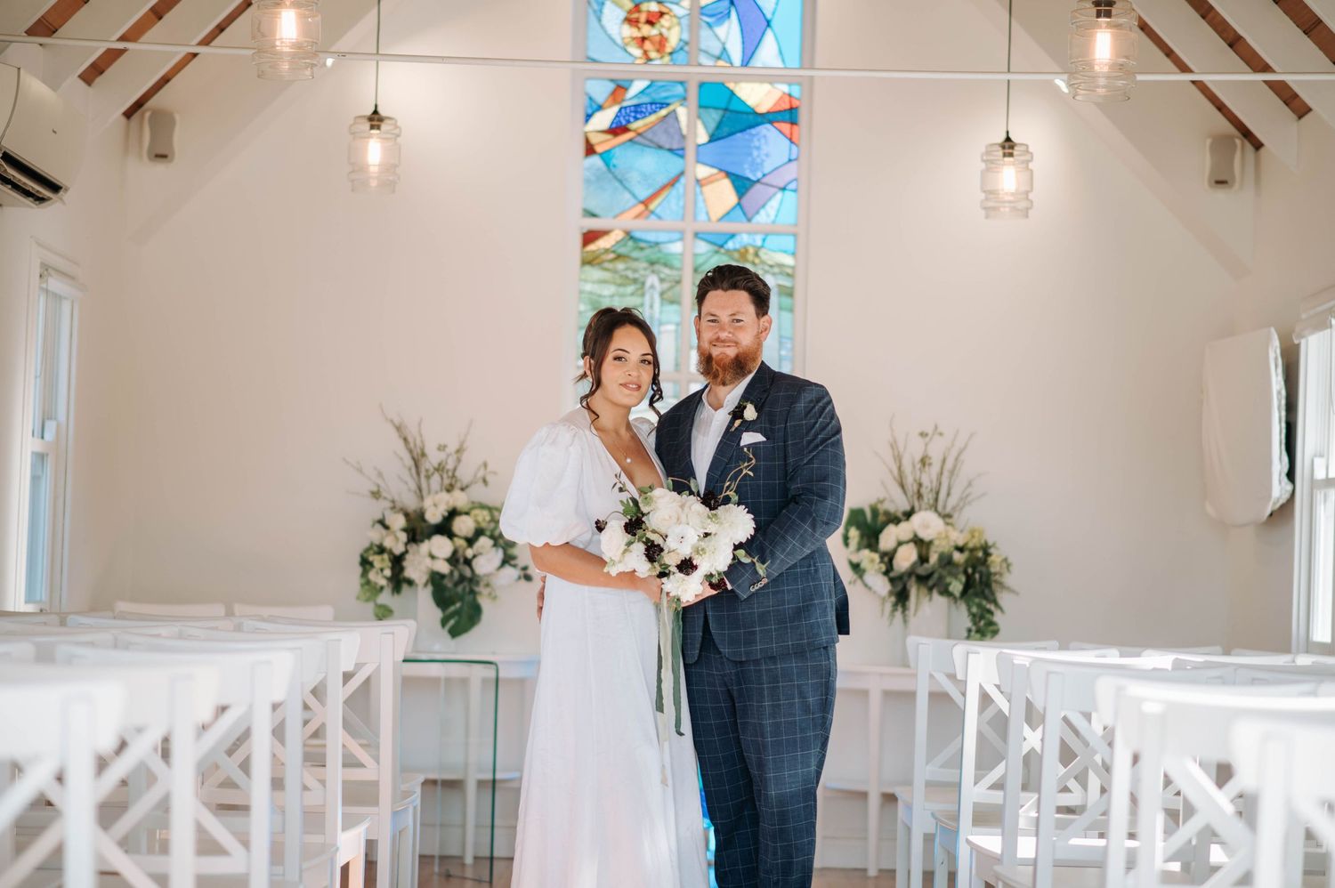 Couple poses in chapel with stained glass windows and hanging pendant lights.
