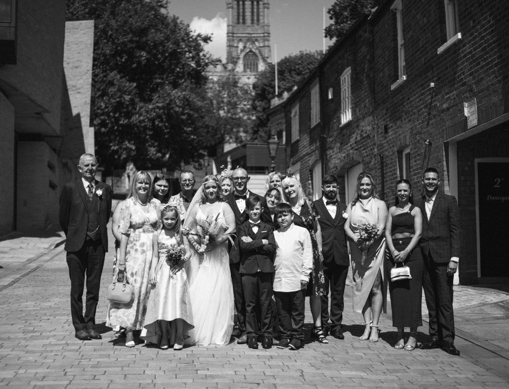 Large wedding party poses together on brick-lined street with church spire visible in background.