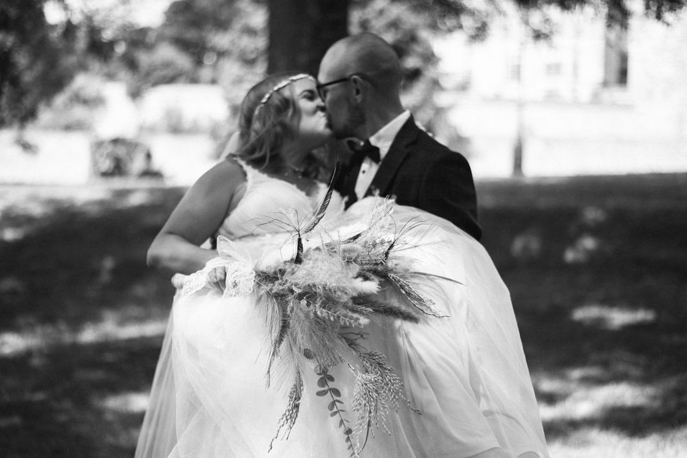 Black and white portrait of newlyweds kissing while holding floral bouquet outdoors.