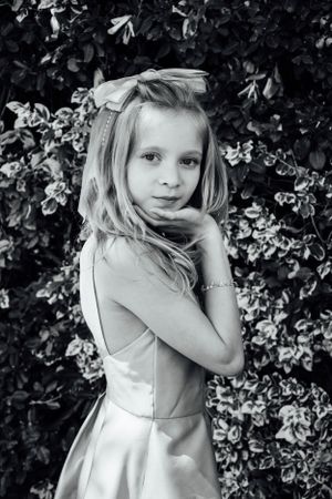 A black and white portrait of a young girl in a formal dress posing against a floral backdrop.