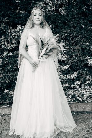 An elegant bride in a flowing white gown and veil holding dried flowers against a garden backdrop.
