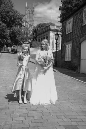 Two people in formal attire pose on a cobblestone street with a historic church tower in the background.