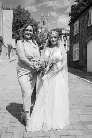 Two people in formal attire stand together on a brick pathway with a church spire visible behind them.