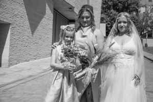 Three people in formal attire pose together holding bouquets against a white wall.
