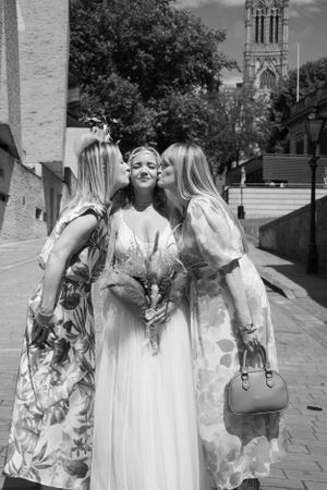 Three friends in floral dresses share an intimate moment on a cobblestone path with a church tower in the background.