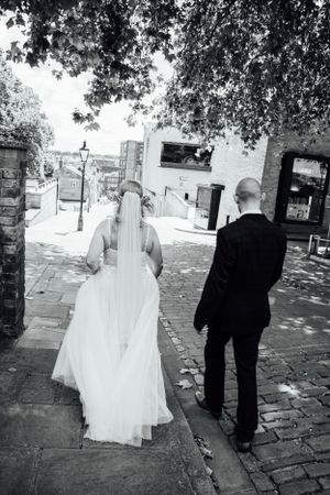 A couple in wedding attire walks along a cobblestone path under shady trees.