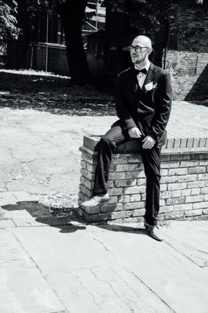 A formal portrait of a person in a bow tie and suit sitting on a brick wall.