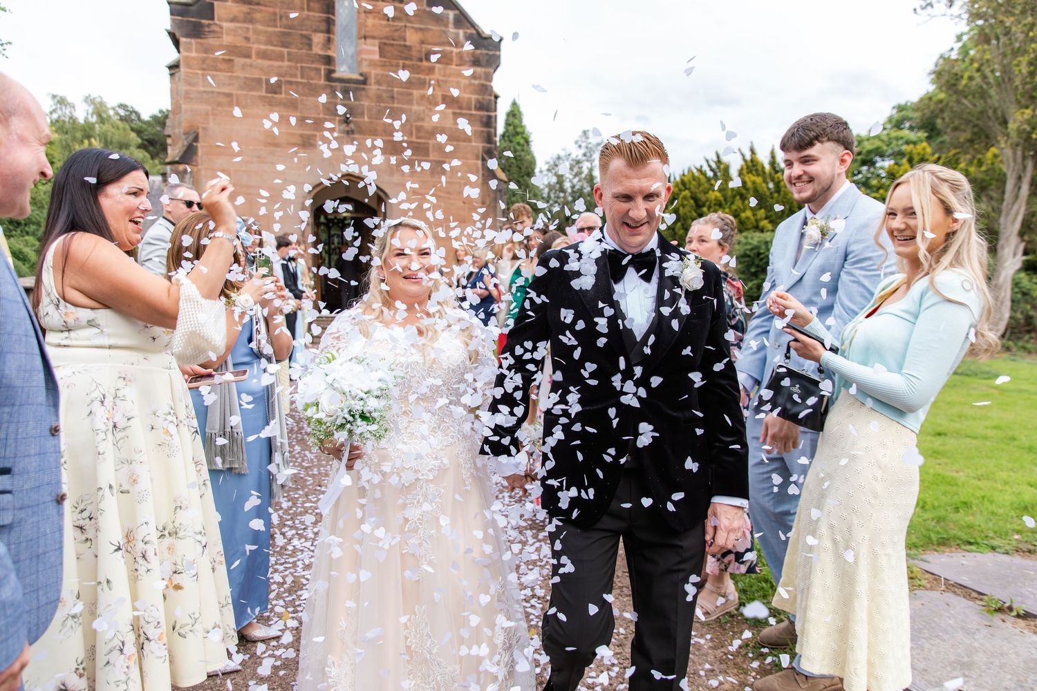 Bride and groom walk through confetti shower surrounded by smiling wedding guests outdoors near brick building.