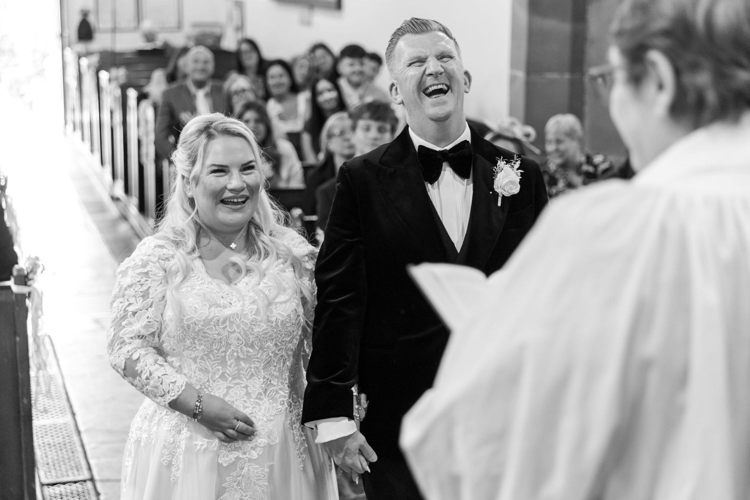 Black and white photo of a laughing bride and groom holding hands at the altar during their church wedding ceremony.