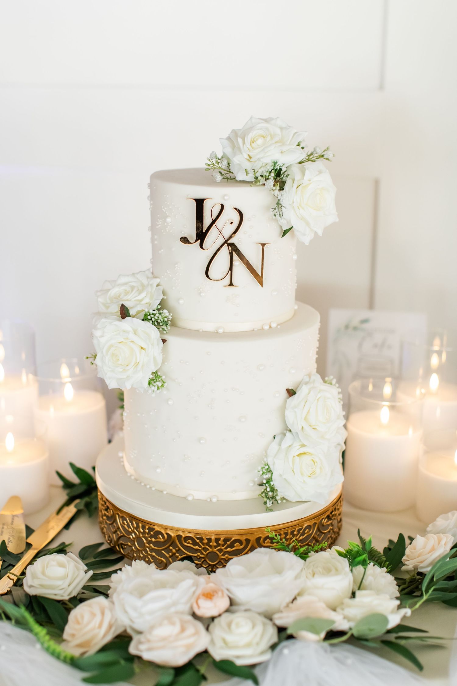 Three-tier white wedding cake with gold monogram, white roses, and candles on ornate gold stand.