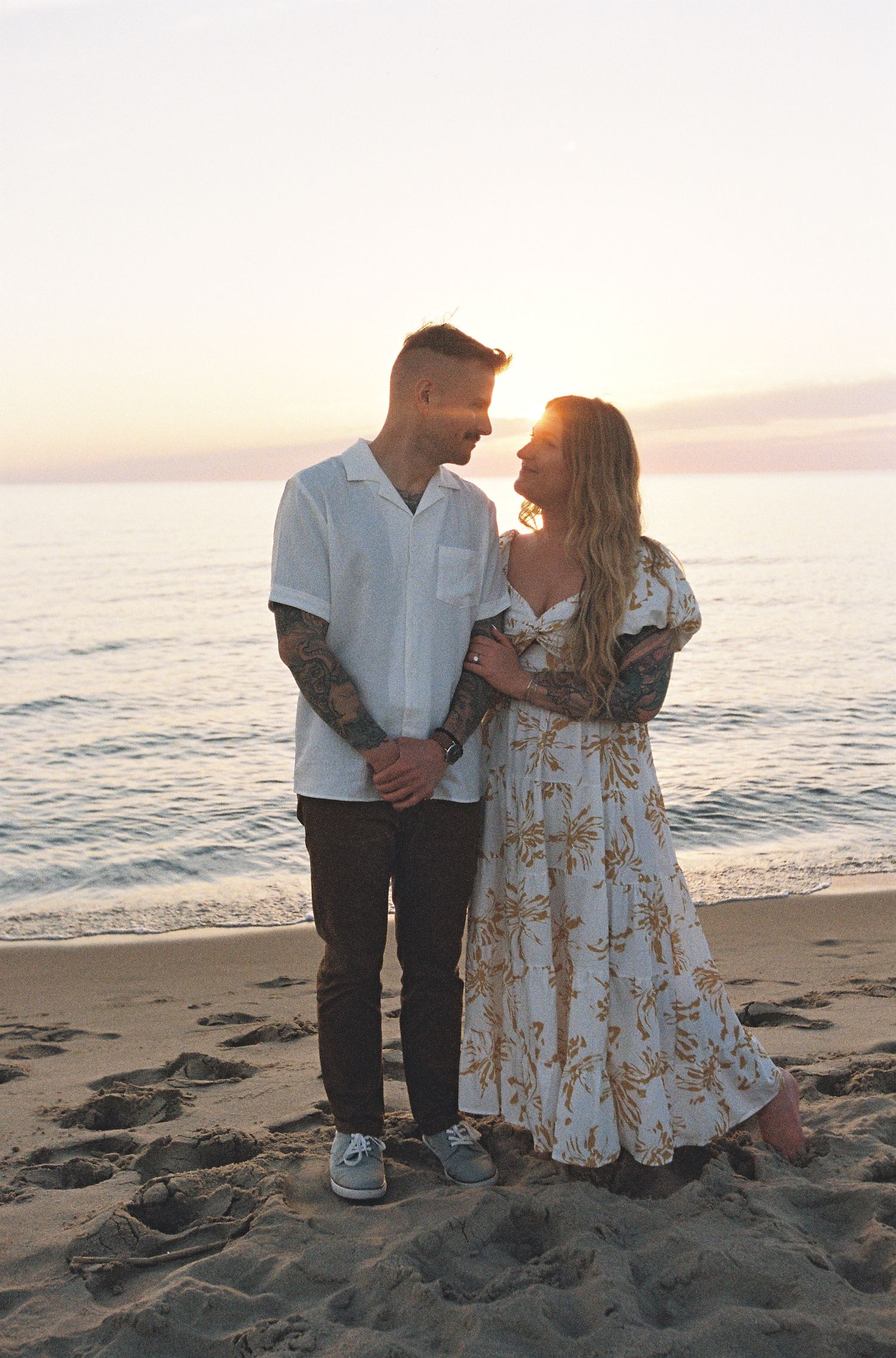 Couple holding hands smiling at each other on sandy beach at golden sunset with calm ocean waves.