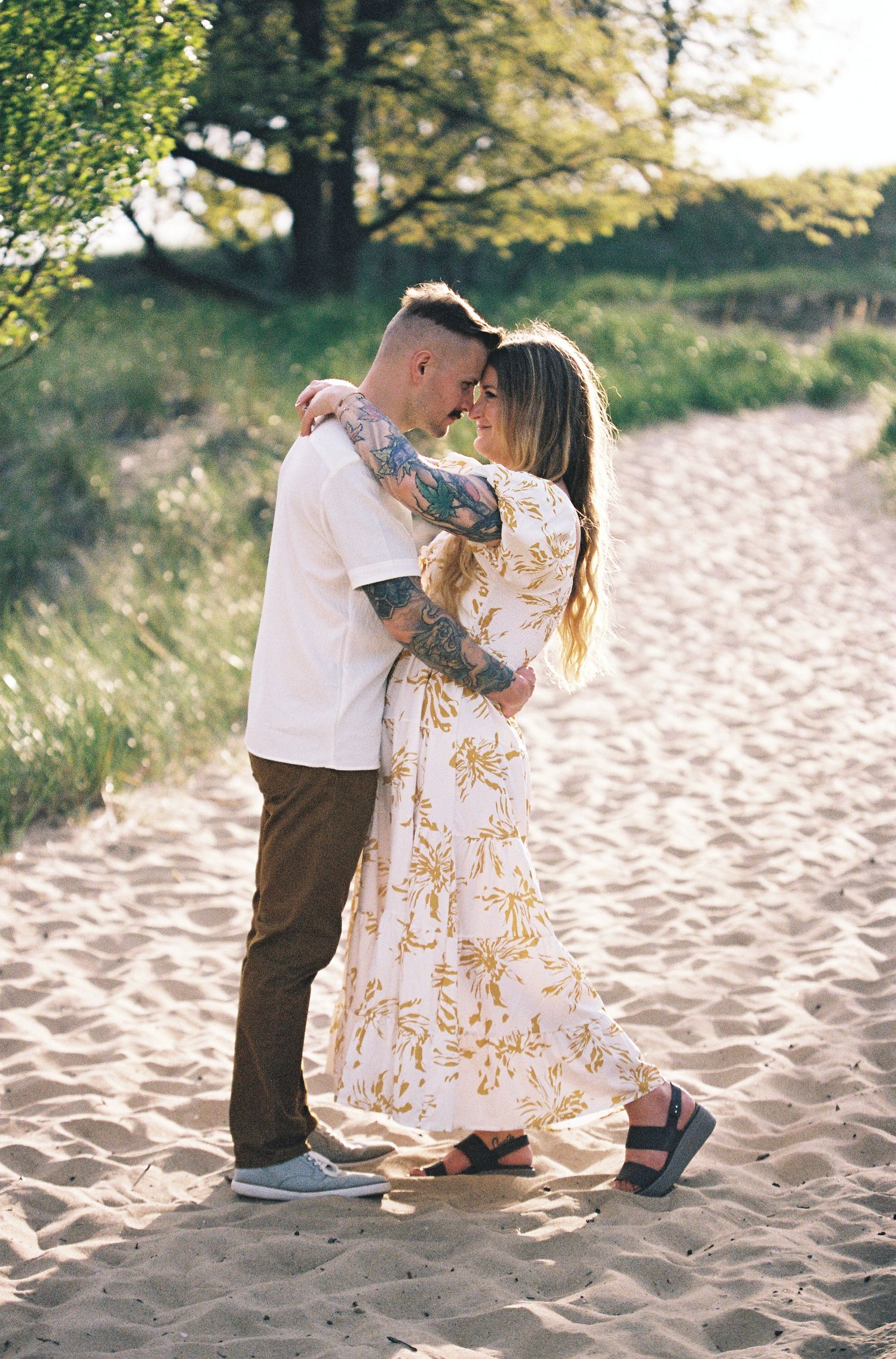 A tattooed couple embraces romantically on a sandy beach path surrounded by lush green trees at golden hour.