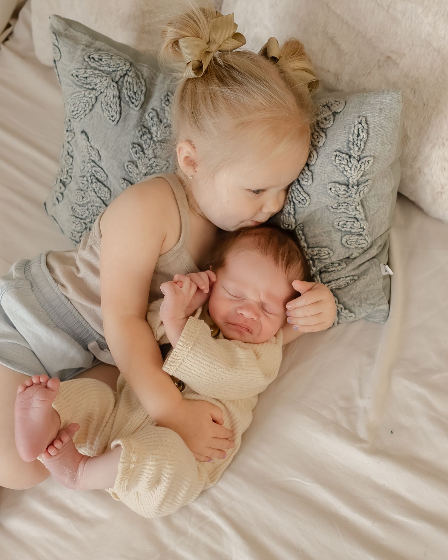Young child tenderly embraces newborn sibling on soft bedding with decorative pillows.