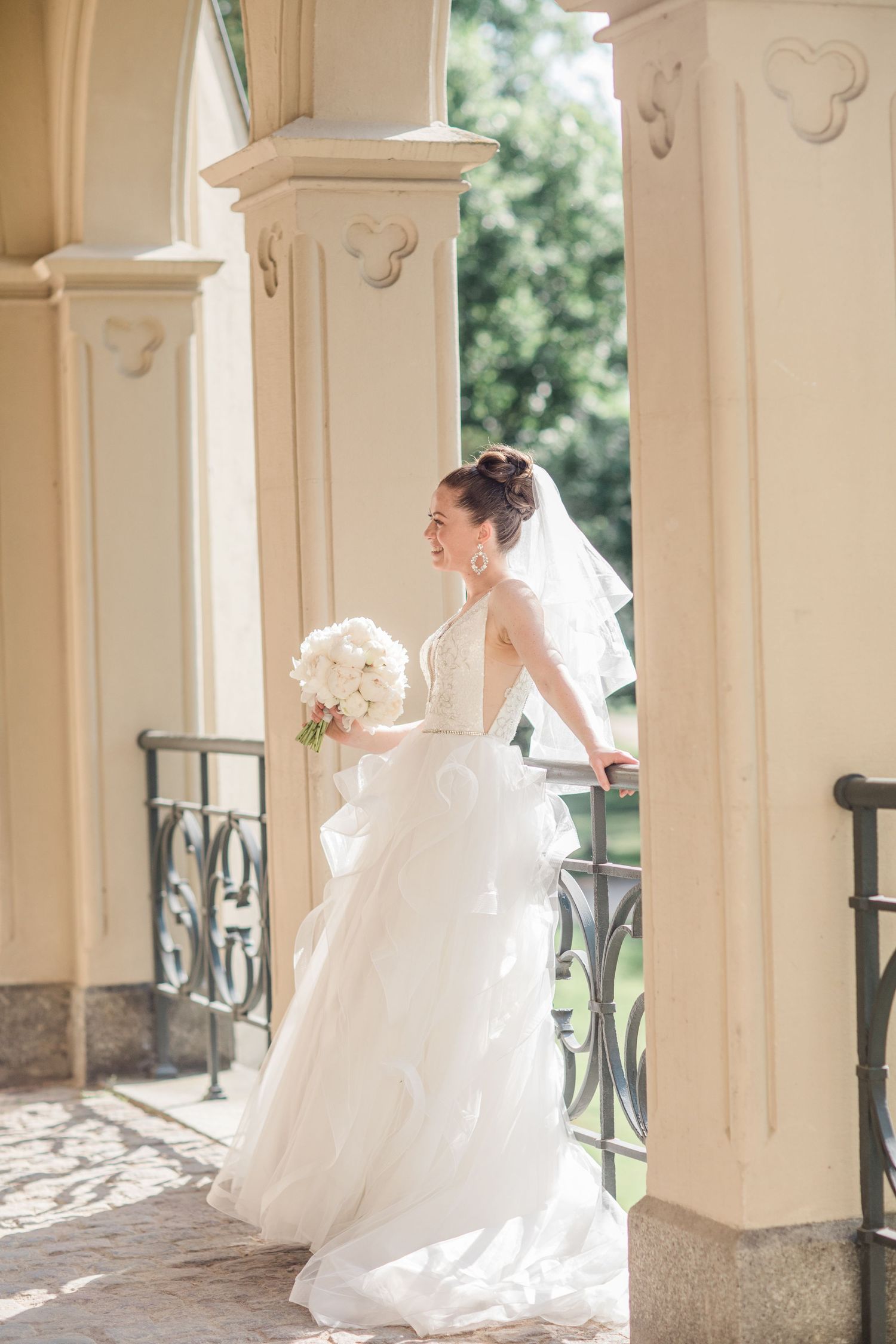 A bride in a flowing white wedding dress with tulle skirt and veil holds a bouquet while standing between elegant columns.