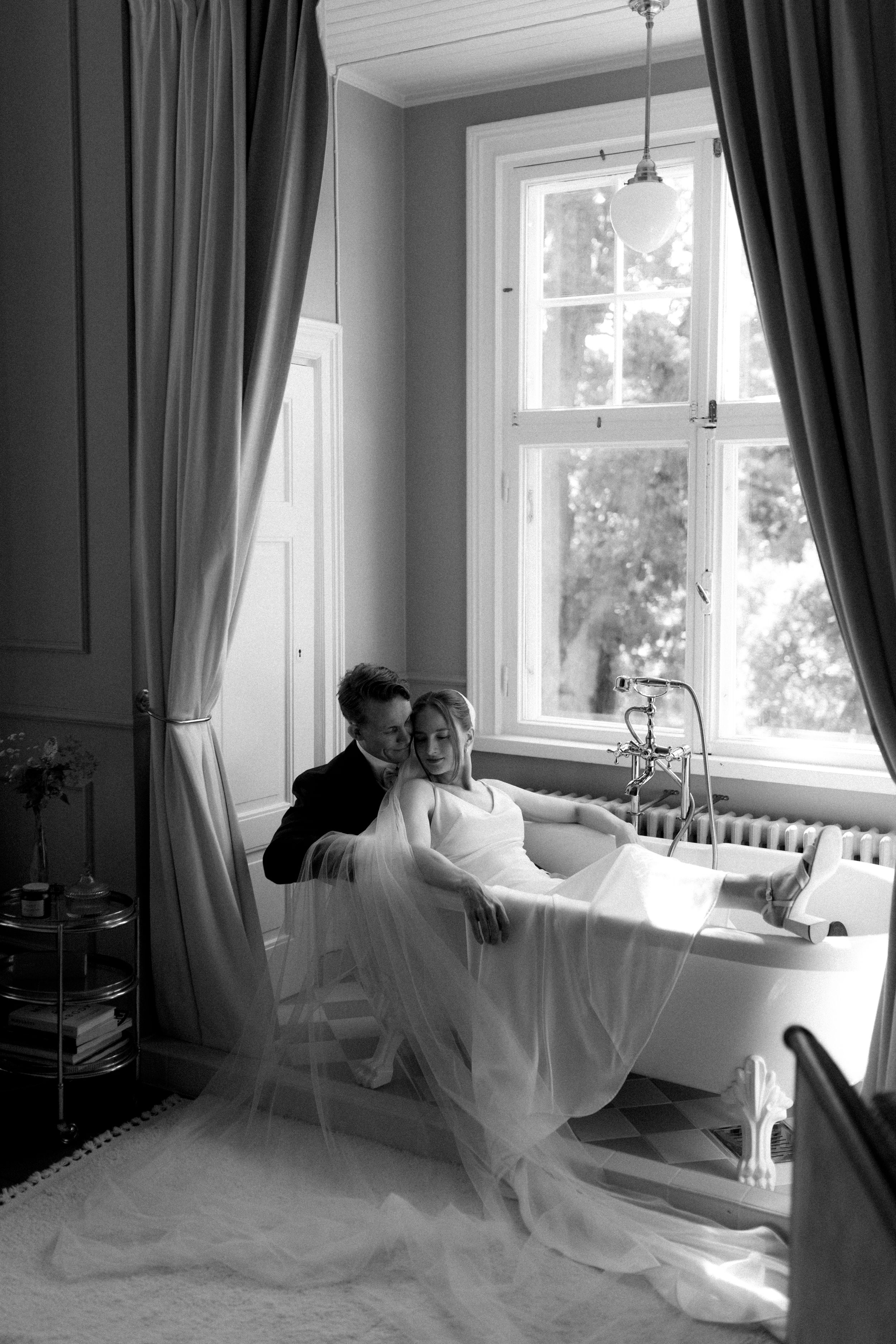 Black and white wedding portrait showing bride and groom sitting in a bath tub in an elegant wedding suite in Billnäs Gård, Finland.