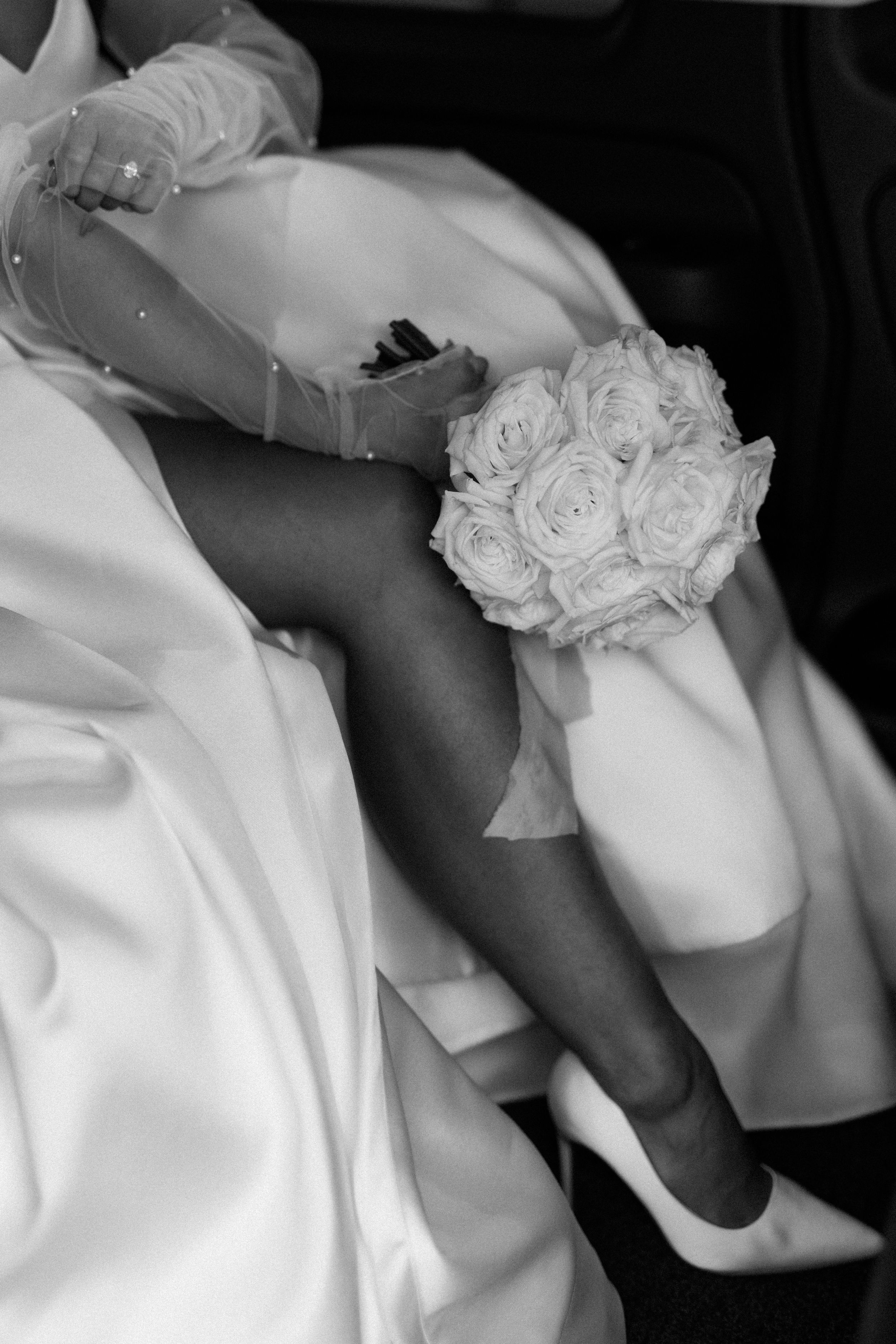A black and white artistic wedding photo showing brides leg and white wedding shoe with white roses adorning a flowing wedding dress fabric.