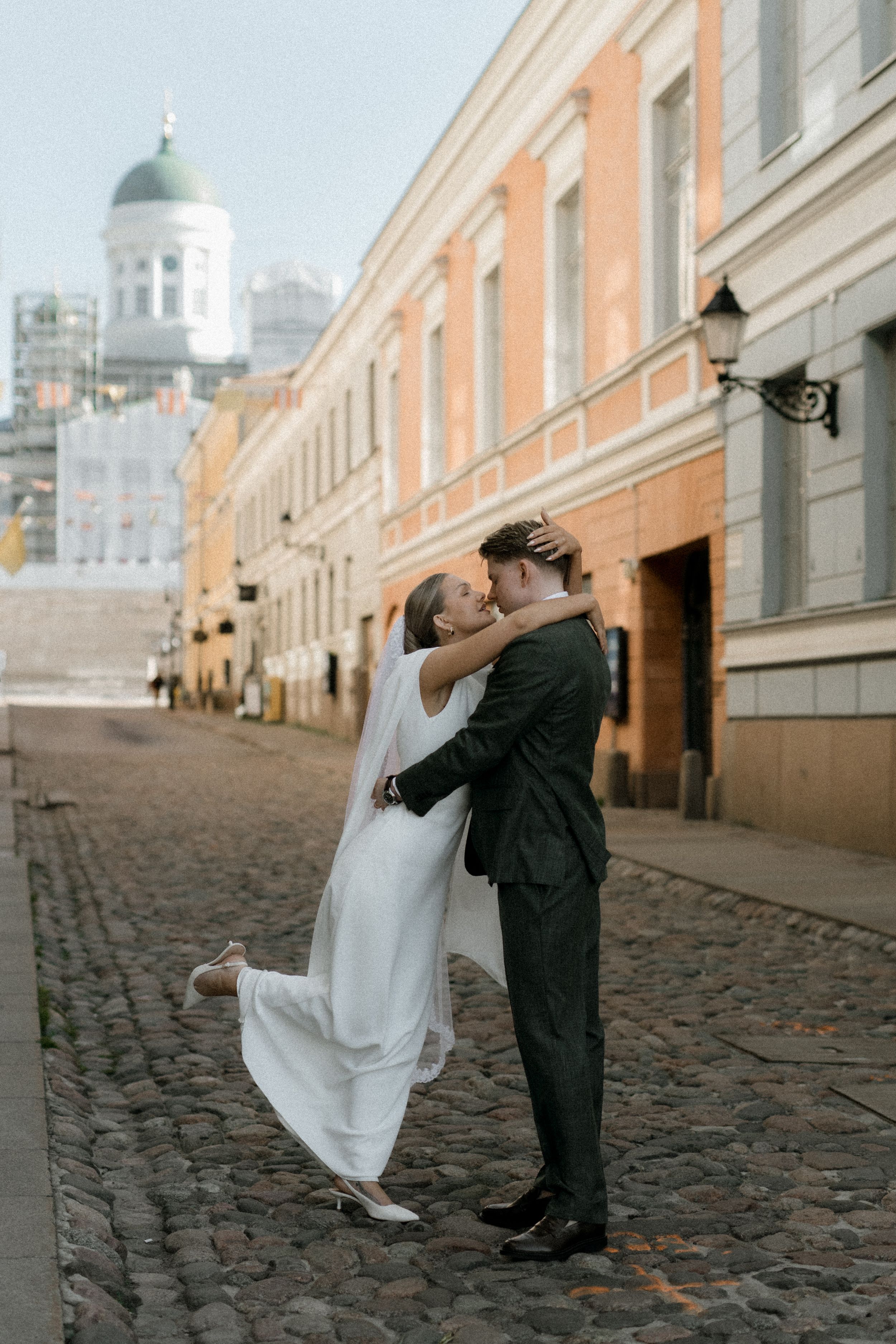 A romantic wedding couple embraces on a cobblestone street in Helsinki with the Tuomiokirkko cathedral dome in background.