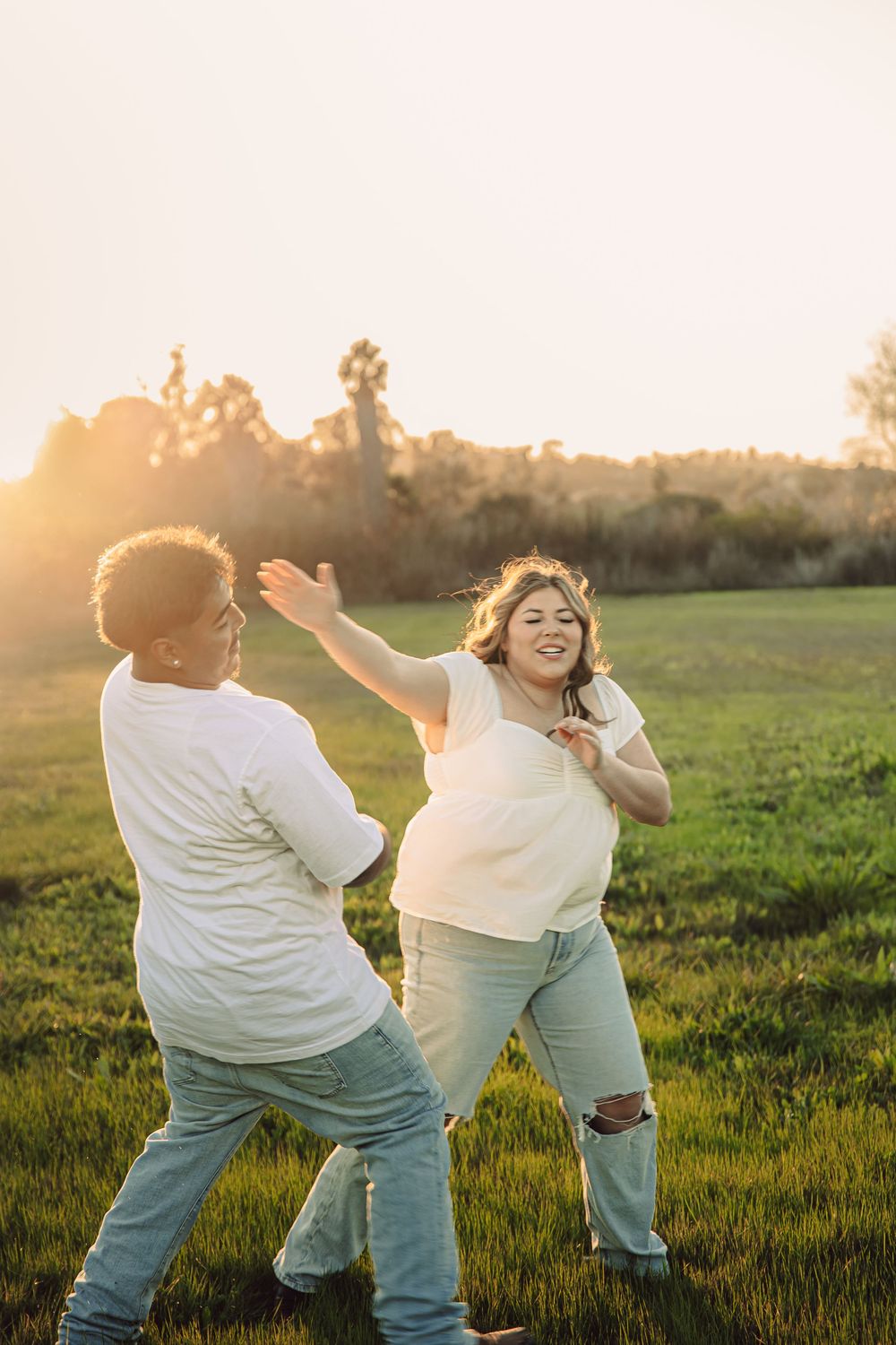 Two people in white shirts and jeans dance playfully together in a sunlit grassy field at golden hour.