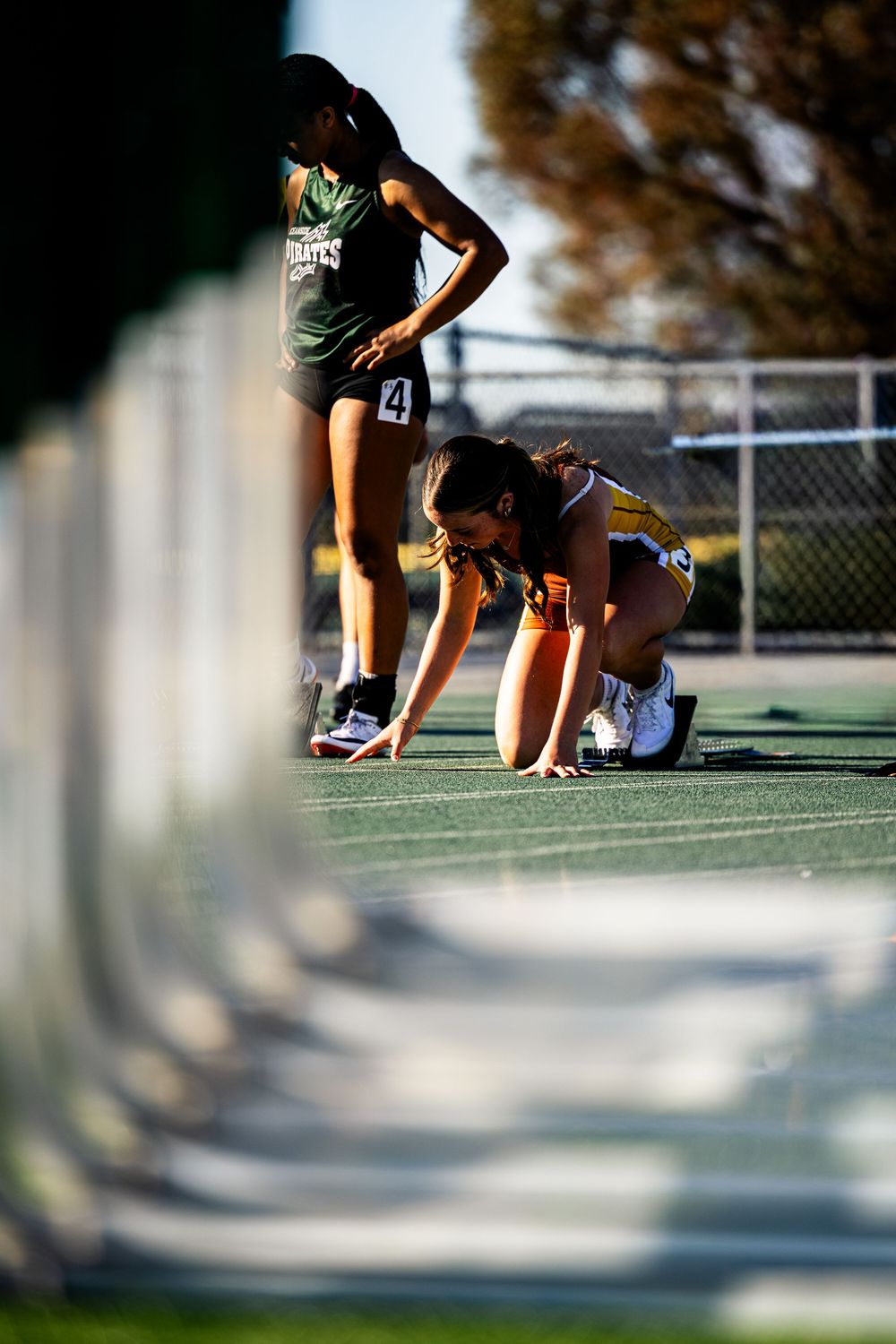 Female track athlete in starting position on green track, with teammate standing behind her at outdoor competition.