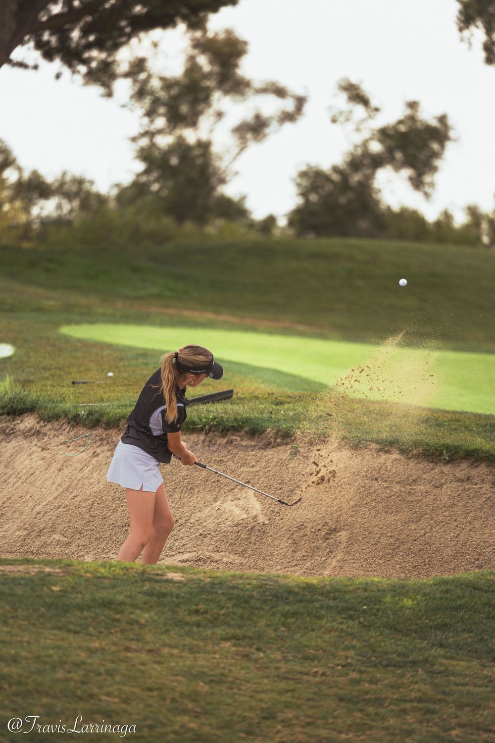 Golfer hitting out of a sand bunker on a scenic golf course at sunset.
