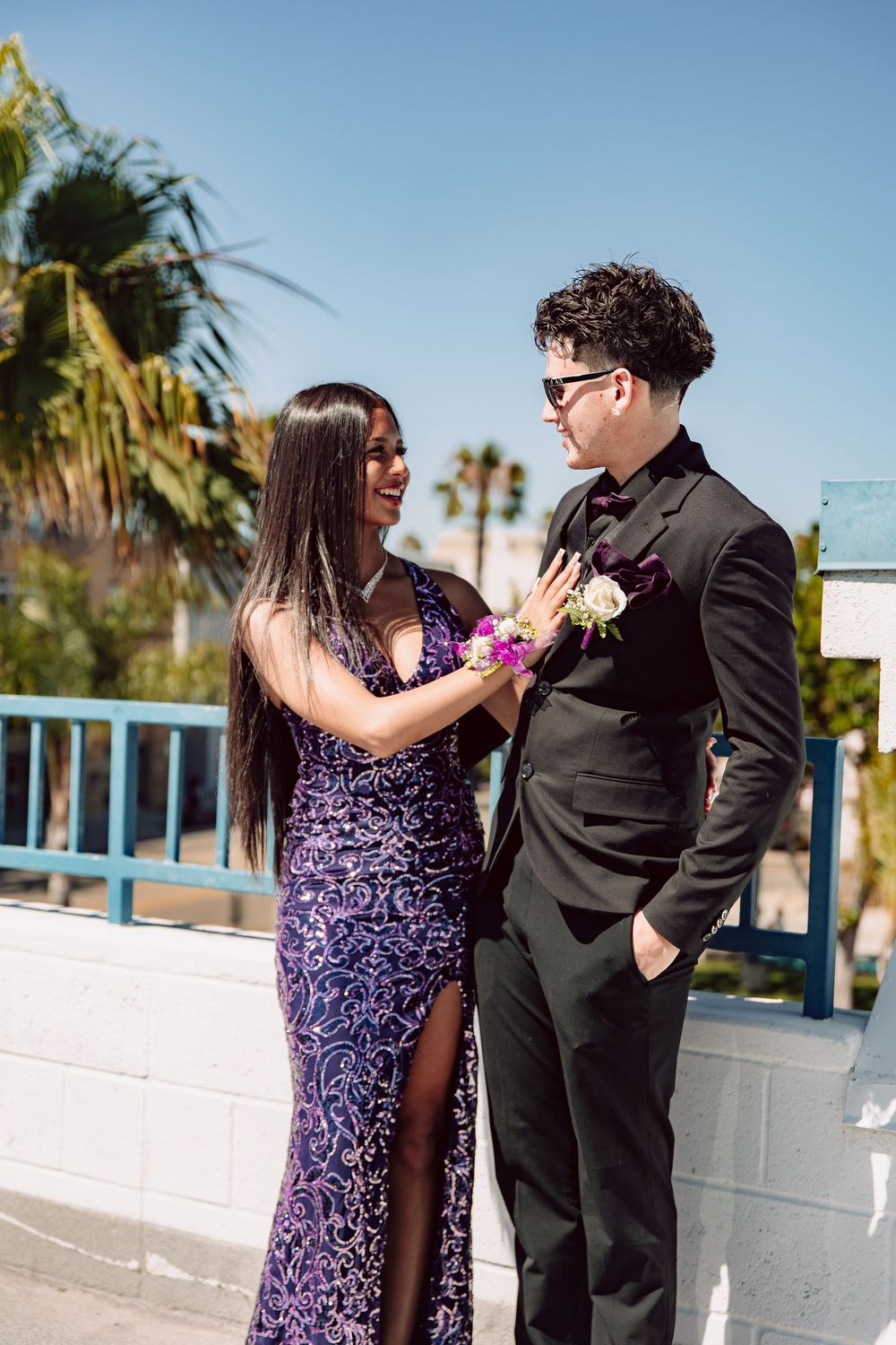 Couple in prom attire, woman in purple sequin gown pins boutonniere on man in black suit outdoors.