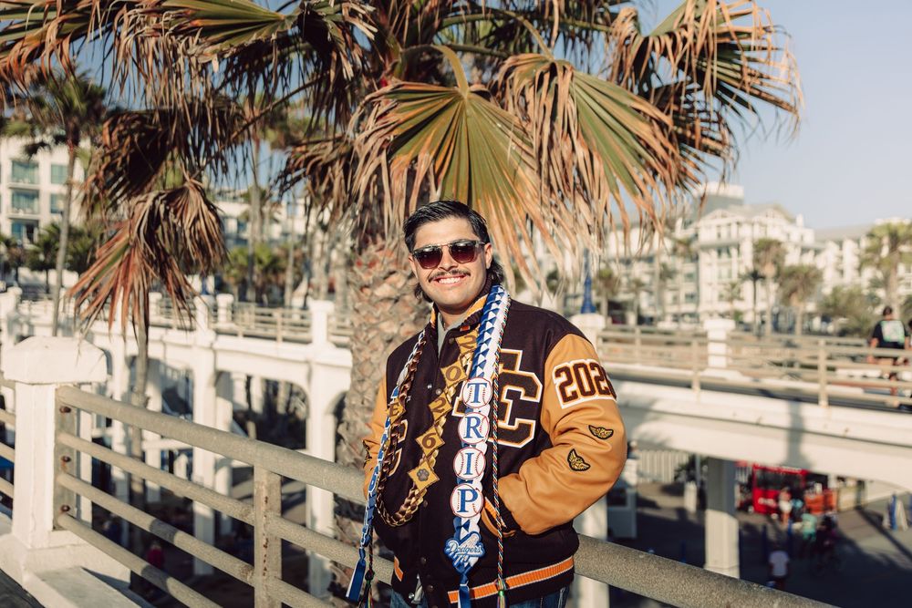 Young man smiling in a varsity jacket and scarf near palm trees on a sunny boardwalk.